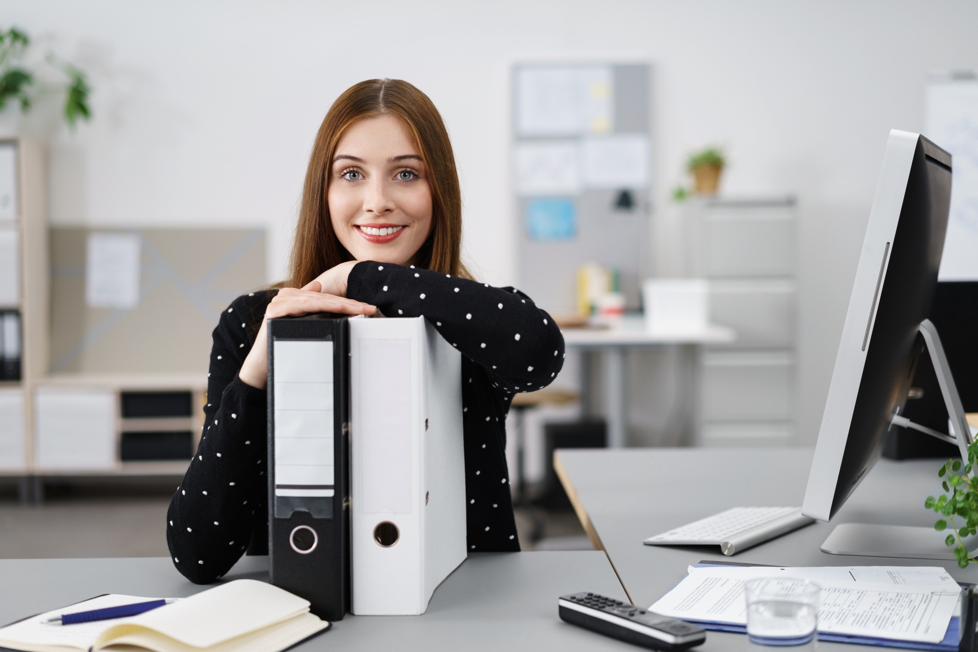 Eine Person steht an einem grauen Schreibtisch in einem hell und steril eingerichteten Büro. Ihre Arme sind an zwei Ordner gelehnt, vor ihr liegt ein Notizbuch und ein Kugelschreiber, neben ihr ein Monitor, Dokumente und eine Fernbedienung. 