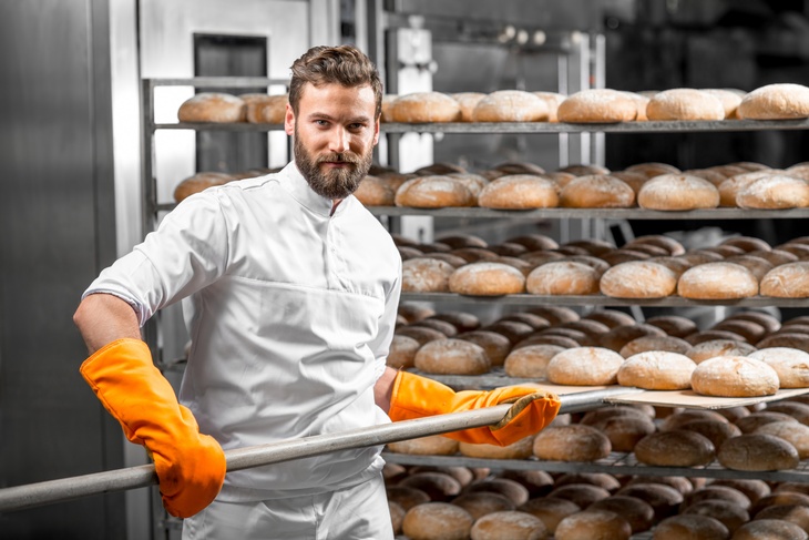 Person mit Bart mit gelben Schutzhandschuhen hievt Bäckerschaufel Brotlaibe aus Regal