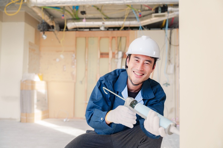 Nahaufnahme einer Person mit einem Helm, die mit einem Gerät zum Spritzen arbeitet. Im Hintergrund eine Baustelle und Kabel. 