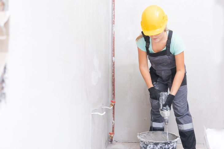 Eine Person rührt mit einem Bohrer eine graue Maße an, die sich in einem Kübel befindet. Sie befindet sich auf einer Baustelle. 