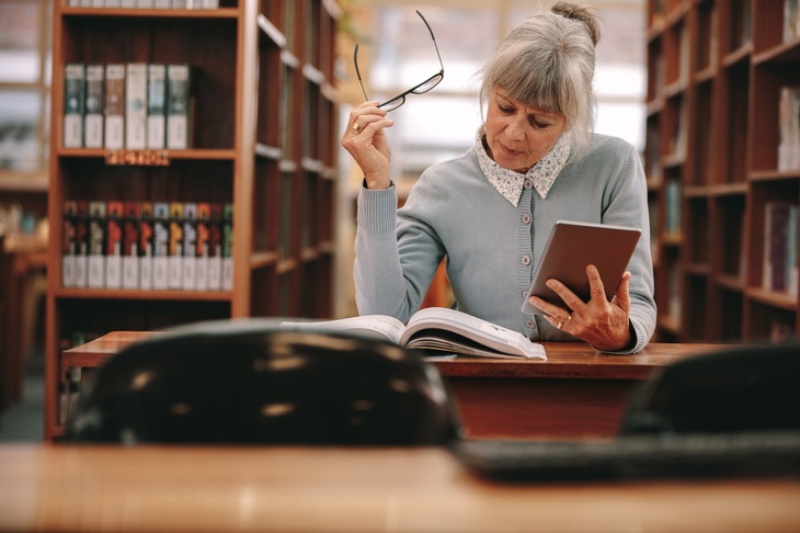 Person sitzt in Bibliothek und blickt auf Buch, hält in einer Hand eine Brille und in der anderen ein Tablet