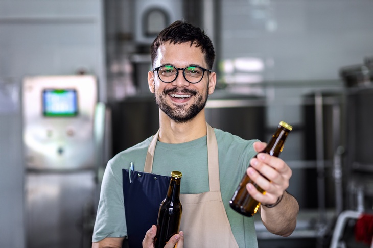 Porträt einer Person mit Brille und Bart, die eine Bierflasche in die Kamera hält. In der rechten Hand hält sie ein Klemmbrett und eine weitere Flasche. Im Hintergrund sind verschiedene Geräte zum Bierbrauen