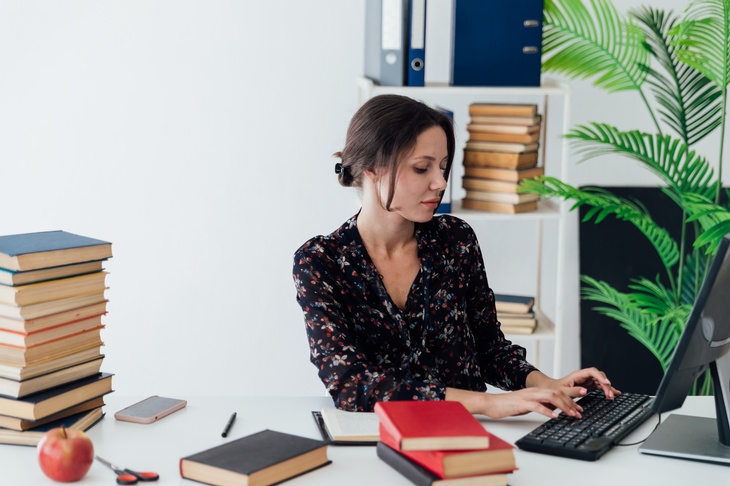 Eine Person sitzt auf einem Schreibtisch, auf dem Stapel von Büchern und ein Apfel liegen. Sie tippt auf der Tatstatur eines Computers. Im Hintergrund sieht man Bücher, Ordner und eine Topfpflanze. Die Stimmung des Bildes ist steril. 