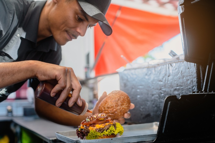 Eine Person hält eine Ketchup-Tube in der rechten Hand und ein Burgerbrötchen in der linken. Sie belegt einen Burger mit einem Fleischlaibchen, Salat und Käse auf einem silbernen Tablett. Im Hintergrund ist ein silberner Tresen und ein orangener Schirm. 