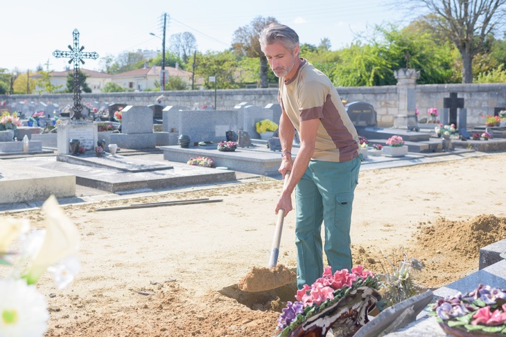 Eine Person gräbt mit einer Schaufel Erde um, sie hebt eine Grube für ein Grab in einem Friedhof aus. Ringsherum sind bunte Blumengestecke und Gräber, man sieht eine Mauer, Häuser und Bäume im Hintergrund des Friedhofs. 