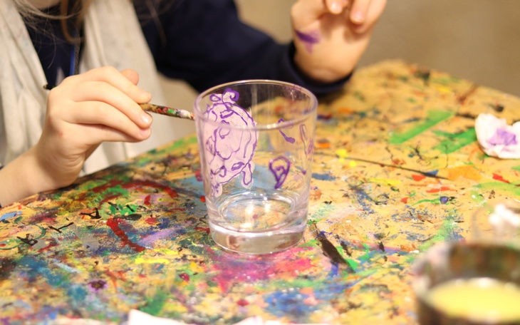 Im Fokus ist ein Glas, das auf einem Tisch steht, der mit Farbe bekleckst ist. Dieses wird mit einem Pinsel verziert, die Büste der  Person, die diesen in der Hand hält, ist in der linken Bildhälfte halb zu sehen.