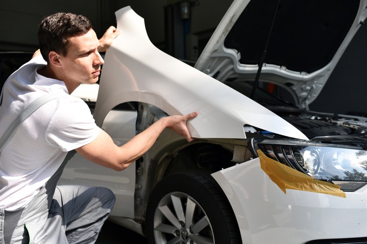 Eine Person in grauer Latzhose ist knapp neben einem weißen Auto, dessen Motorhaube geöffnet ist. Die Person hält ein weißes Teil auf das Auto. Beim vorderen Scheinwerfer des Autos ist ein braunes Klebeband