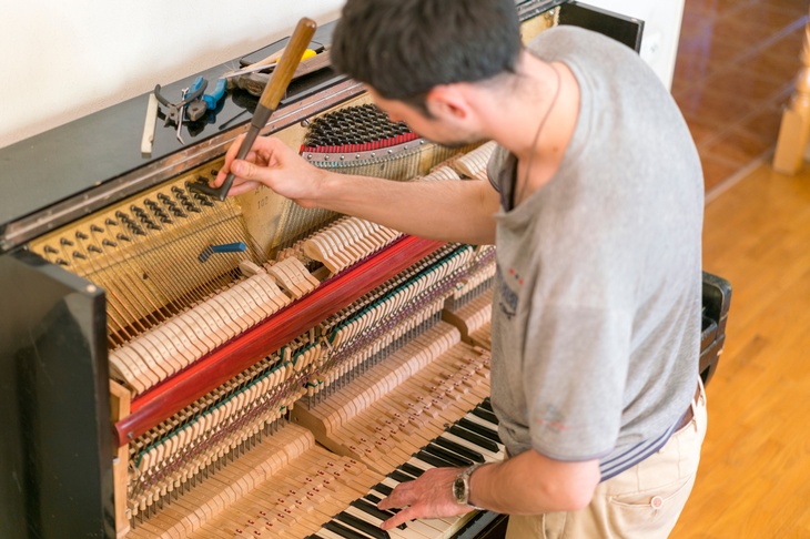 Eine Person steht an einem Klavier. Der Bereich oberhalb eines Klaviers, wo die schwarz-weißen Tasten sind, ist geöffnet, wo verschiedene Holztasten und Drähte sind. Die Person hält ein Werkzeug auf einen Draht