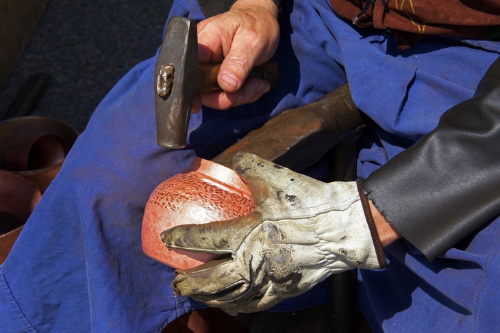 Nahaufnahme von Händen einer Person, in der linken Hand hält sie eine braun-rote, rundliche Form mit einem Arbeitshandschuh. In der rechten Hand hält sie einen Hammer, mit der sie das Objekt bearbeitet. 