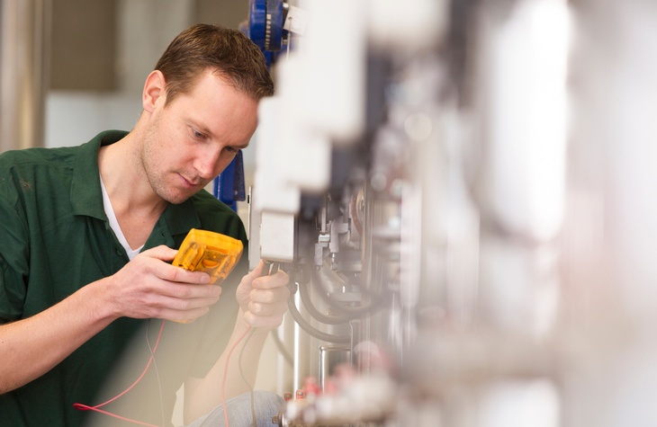 Person mit kurzen Haaren und grünem Poloshirt misst mit einem Gerät einen Wert einer industriellen Anlage