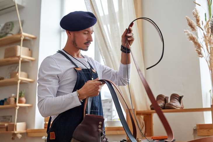 Seitliche Aufnahme einer stehenden Person, die in den Händen lange, schmale Lederriemen hält und darauf blickt. Hinter ihr steht auf einem Regal ein Paar Lederschuhe. Im Vordergrund ist ebenfalls ein Lederschuh.