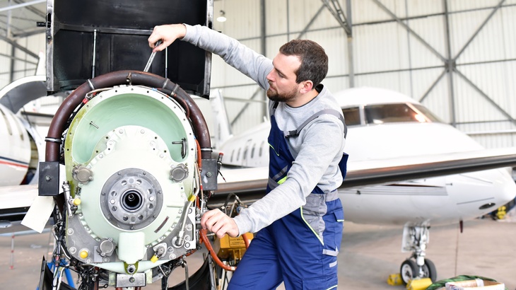 Person in blauer Arbeitslatzhose schraubt an Triebwerk eines Flugzeuges in Hangar, im Hintergrund verschwommen geparktes Flugzeug