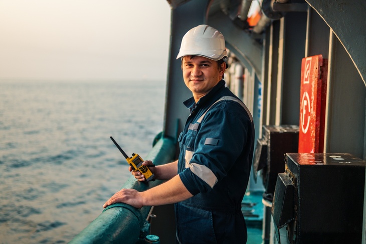 Person in Arbeitsoverall mit weißem Helm steht an Reling eines Bootes und hält Walkie Talkie in der Hand, im Hintergrund verschwommen Meer