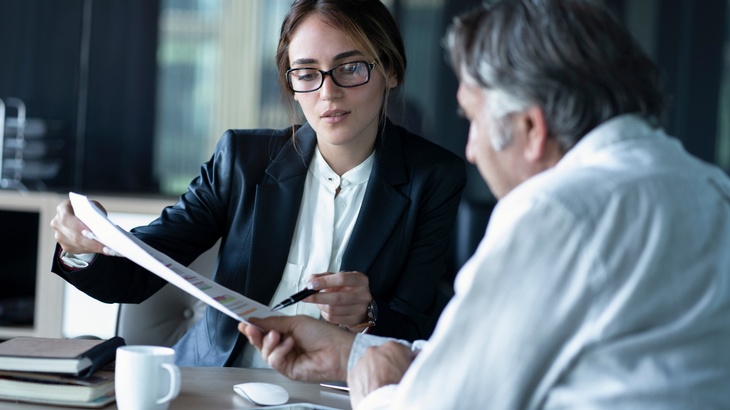 Person in Businesskleidung und Brille erklärt einer weiteren Person Unterlagen