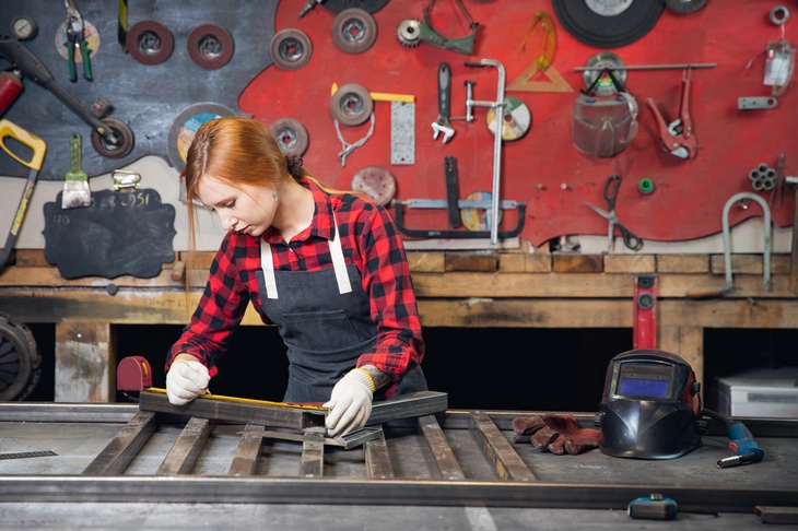 Frontalaufnahme einer an einem Tisch stehenden Person mit Handschuhen. Vor ihr liegt eine angewinkelte, rechteckige Metallkonstruktion, auf die die Person ein Maßband hält. Rechts neben ihr liegt ein Gesichtshelm