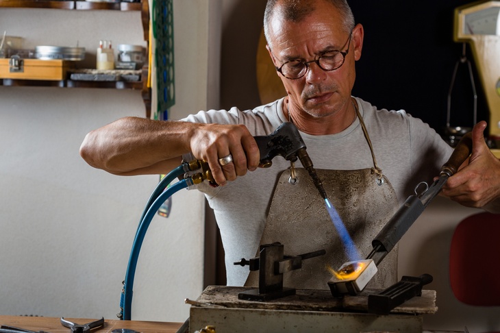 Frontalansicht einer Person mit Schürze, die in der rechten Hand ein längliches Gerät hält, aus dem vorne eine Flamme austritt. Mit der linken Hand hält sie ein quadratisches Metallstück mit einer langen Zange, auf die die Flamme trifft