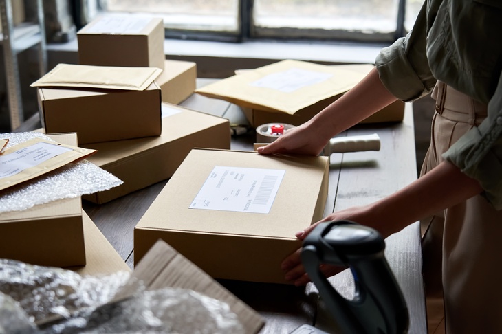 Auf einem Tisch liegen mehrere Pakete, Kuverts sowie durchsichtige Folie. Eine Person steht seitlich vor dem Tisch und hat ihre beiden Händen auf einem Paket