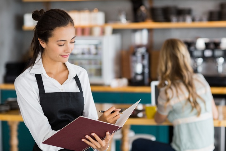 Person mit dunklen geschlossenen Haaren, weißer Bluse und Arbeitsschürze steht in einem Lokal vor einer Theke, im Hintergrund sitzt eine Person mit einem Laptop bei einem Tisch