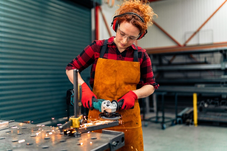 Eine Person mit Gehörschutz, Schutzbrille, Schürze und Handschuhen steht an einem Metalltisch. Mit beiden Händen hält sie ein Gerät, dessen Ende eine Schleifscheibe hat. Das Gerät berührt ein längliches, am Tisch befestigtes Objekt