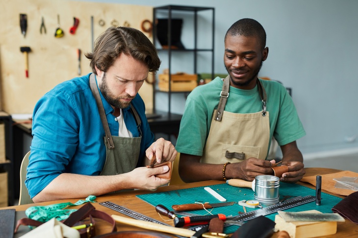 Zwei Personen sitzen auf einem Werktisch, eine davon bearbeitet eine Geldbörse aus Leder, die andere sieht zu. Am Tisch liegen Werkzeuge und Unterlagen zur Verarbeitung von Lederwaren. Kulisse ist eine Werkstatt, an den Wänden hängen Werkzeuge. 