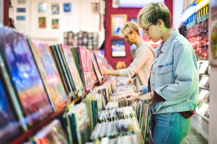 Person in Jeansjacke steht an Regal voller Schallplatten und durchblättert diese, im Hintergrund verschwommen weitere Person beim Stöbern