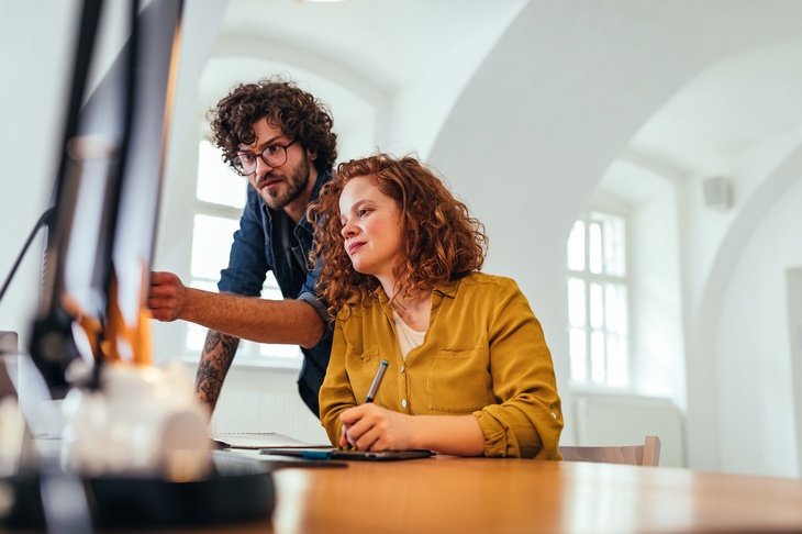 Zwei Personen vor Computerbildschirm, eine Person sitzend, die andere stehend auf Monitor deutend