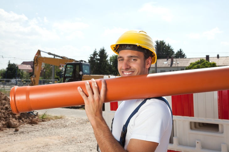 Eine Person hält ein Rohr aus PVC in der Hand. Sue steht auf einer Baustelle, im Hintergrund sieht man einen Kran. 