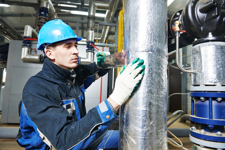 Das Bild zeigt einen Arbeiter in blauer Schutzkleidung und einem blauen Helm, der an einem großen, mit silberner Isolierung umwickelten Rohr arbeitet.  Im Hintergrund sind weitere Rohre zu sehen.